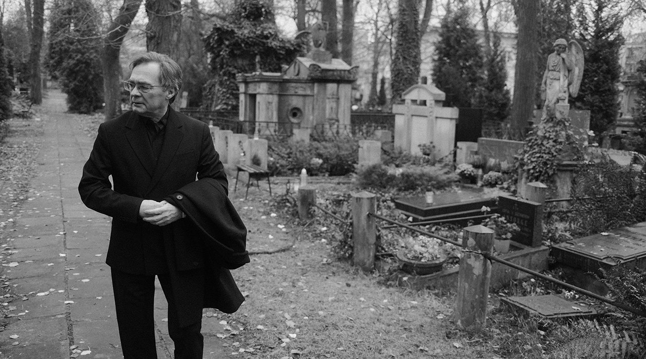 Mistrz idzie alejką cmentarną i patrzy na prawo - baner historii pożegnań